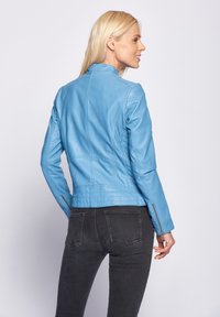 Blaues Lederjackett mit einer taillierten Passform, minimalistischem Kragen und Reißverschlussärmeln. Dunkle Jeans vervollständigen das Outfit, das von hinten gezeigt wird.