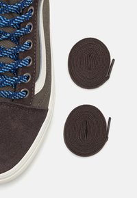 Basket haute en daim marron avec des lacets bleus et noirs, une bande blanche sur le côté, et deux lacets marron enroulés à côté sur un fond blanc.