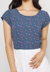 Blå blommig topp med korta ärmar, med rundad halsringning och små rosa och vita blommönster, tillverkad av lättviktsmaterial.