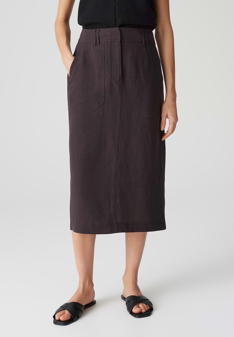 Femme portant un haut noir sans manches, une jupe midi marron foncé avec poches avant et des sandales plates noires à bout ouvert, debout sur un fond uni.