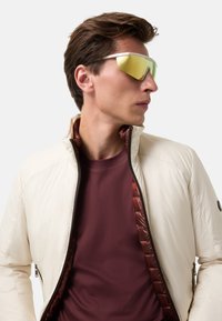 Gepolsterten cremefarbene Jacke mit hohem Kragen, kontrastierendem kastanienbraunen Innenfutter und einem Reißverschluss vorne. Model trägt gelbgetönte Sonnenbrille über einem kastanienbraunen Hemd.