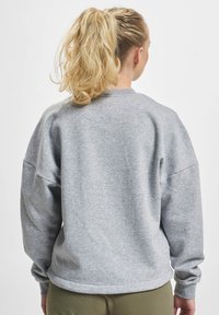 Szürke pulóver laza szabással, bordás mandzsettákkal és kerek nyakkivágással. Simogatós anyag, minimális részletezéssel, hátulról nézve.