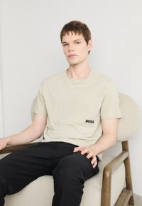 Beige bomullströja med korta ärmar, som har en svart broderad "BOSS"-logga på vänster sida, tillsammans med svarta byxor.