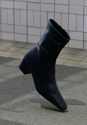 Botte noire en cuir avec talon bloc, posée sur une surface en asphalte texturée près d'un mur carrelé.
