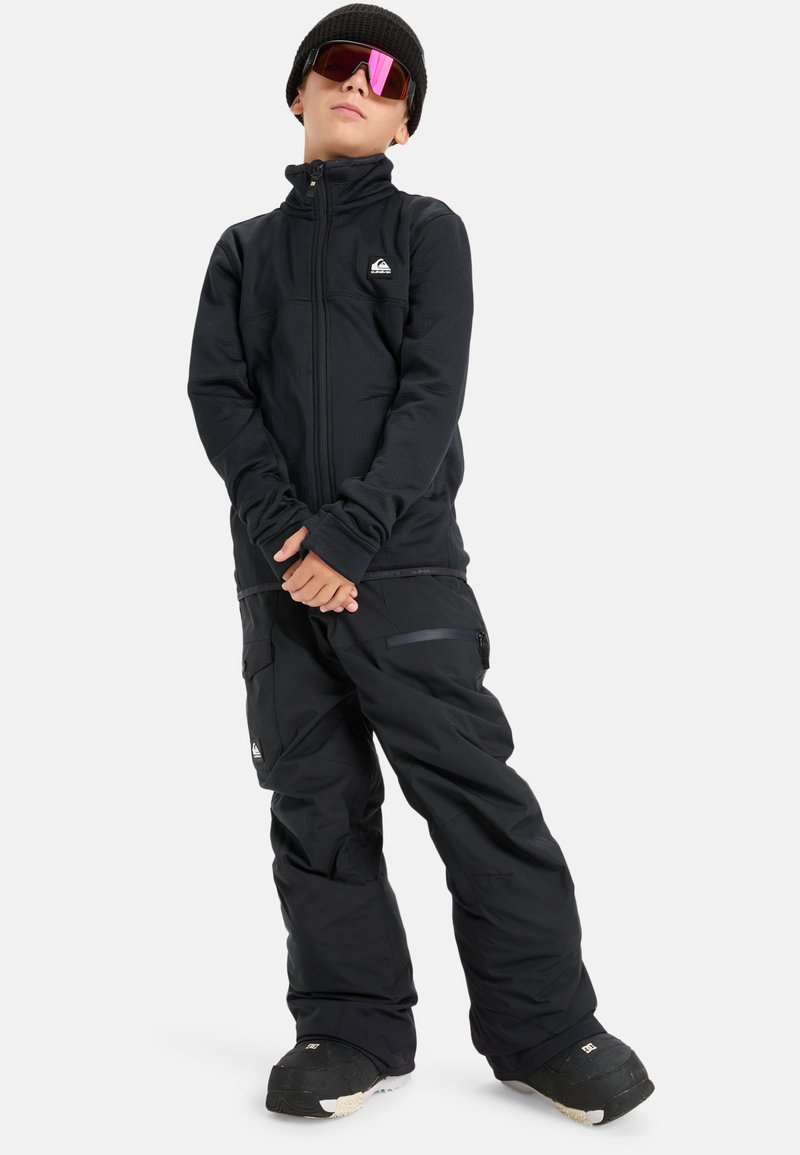 Czarna kurtka narciarska z wysokim kołnierzem i zamkiem, w połączeniu z czarnymi spodniami narciarskimi. Charakteryzuje się fakturowanym materiałem i warstwowym wyglądem. Czarna czapka beanie i różowe okulary przeciwsłoneczne.