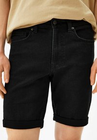 Mustat farkkushortsit, joissa on käännetyt lahkeet, edessä nappikiinnitys ja vyölenkit, kädet lantiolla, vähäisellä yksityiskohdalla.