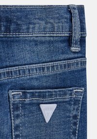 Jeans in denim blu con un logo bianco triangolare sulla tasca posteriore, caratterizzati da cuciture a contrasto e una texture liscia.