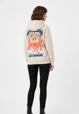 Beiger Hoodie aus weichem Material mit einem bunten Blumenmotiv auf dem Rücken, mit Kapuze und lässiger Passform. Schwarze Slim-Jeans getragen.