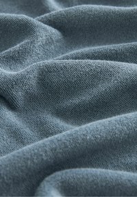 Pehmeä, teksturoitu kangas hillityssä sinisessä sävyssä, jossa on sileä, hieman pörröinen pinta ja näkyviä ryppyjä ja taitoksia.