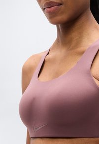 Αθλητικό σουτιέν Nike σε απαλό μωβ, με απαλή υφή, λαιμόκοψη scoop, φαρδιές τιράντες και διακριτικό λογότυπο στην αριστερή πλευρά.