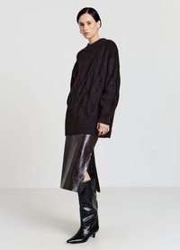 Dunkelbrauner Strickpullover mit Zopfmuster und hohem Kragen, kombiniert mit einem glänzenden bordeauxroten Midirock und schwarzen kniehohen Stiefeln mit spitzer Spitze.