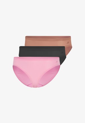 Frau steht barfuß, trägt einen rosa Sport-BH und passende Bikini-Unterwäsche, schaut leicht nach links vor einem weißen Hintergrund.