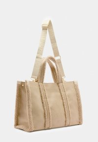 Beige handbag with plush side accents and textured surface. Features dual handles and a detachable strap with embossed branding.