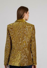 Gouden blazer met pailletten en een textuur met golvende patronen, met lange mouwen en een gekarteld kraag, van achteren weergegeven op een model.