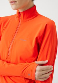 Lys orange treningsgenser med høy krage og halvmeter glidelås, med lange ermer og en tettsittende design. Lite logo på brystet.