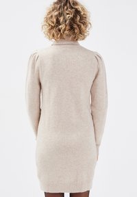 Vestido de punto beige con mangas largas, cuello alto y hombros acampanados. Textura suave y bordes acanalados en los puños y el dobladillo.