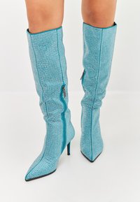 Bocanci turquoise, până la genunchi, acoperiți cu mici strasuri, având nasul ascuțit și un fermoar pe lateral pentru o îmbrăcare ușoară. Incluzând toc stiletto.