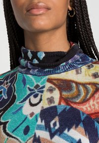 Col roulé coloré à motifs mêlant dessins floraux et géométriques, avec un col côtelé aux tons bleus et noirs.