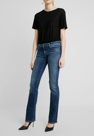 Frau trägt ein schwarzes kurzärmeliges Oberteil, blaue Jeans mit geradem Bein, schwarze spitze High Heels und große Creolen vor einem schlichten grauen Hintergrund.