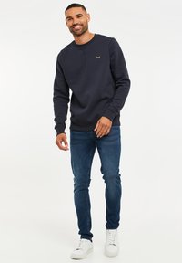Marineblaue Sweatshirt mit Rundkragen und gerippten Bündchen, versehen mit einem kleinen goldenen Logo. Kombiniert mit schmal geschnittenen blauen Jeans und weißen Sneakern.