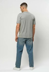 Mann som står med ryggen til iført en grå kortermet t-skjorte, blå jeans, hvite joggesko og et sølvkjede.