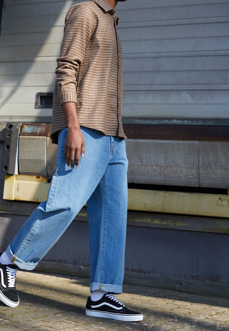 Ruskea ruudullinen paita, vaaleansiniset farkkuhousut ja mustat lenkkarit. Paidassa on kaulus ja pitkät hihat; housut ovat löysät.