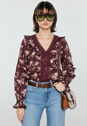 Kobieta nosząca duże żółto-podobne okulary przeciwsłoneczne, bordową bluzkę z falbanami w kwiaty, niebieskie jeansy z brązowym paskiem oraz niosąca dwukolorową torebkę.