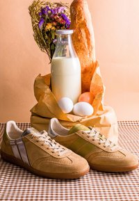 Paire de baskets en daim beige sur une surface à carreaux, derrière un sac en papier contenant une baguette, une bouteille de lait, trois œufs et un bouquet de fleurs séchées.