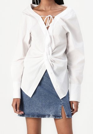 Femme portant un chemisier blanc à manches longues avec un détail nœud à l'avant et une mini jupe en jean bleu avec une fente à l'avant.