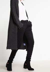 Svarta texturerade byxor som är kombinerade med en vit skjorta och en lång, mörkgrå, stickad kofta. Byxorna har synliga sömmar och detaljer.