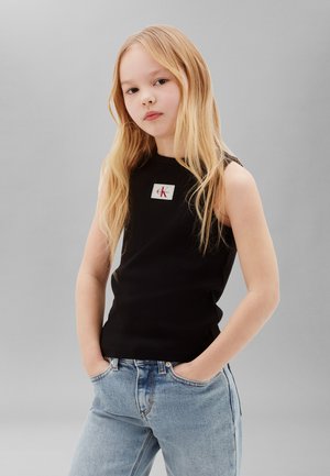 Junges Mädchen mit langen blonden Haaren, das ein schwarzes ärmelloses Top mit Logo und hellblaue Jeans trägt, steht mit den Händen in den Taschen.