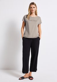 Grijze kortekmouwtop met bedrukte tekst, gecombineerd met zwarte wijde broek en zwarte slide sandalen. Model staat met handen in de zakken.