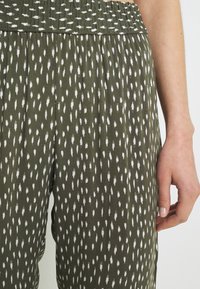 Zielone spodnie w wzory mają marszczony pas, teksturowany materiał oraz wzór z małymi białymi plamkami przypominającymi pociągnięcia pędzlem.