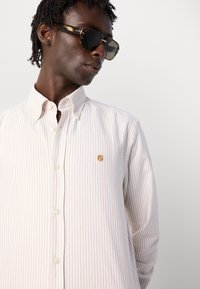 Camicia a maniche lunghe, con bottoni, di un beige chiaro con sottili righe verticali. Presenta un piccolo logo dorato sul petto e un colletto classico.