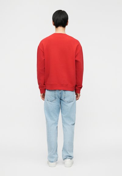 Άνδρας με κοντά μαύρα μαλλιά στέκεται με την πλάτη, φοράει κόκκινο φούτερ, ανοιχτόχρωμο μπλε τζιν και λευκά αθλητικά παπούτσια σε απλό λευκό φόντο.