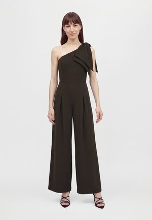 Une femme porte une combinaison noire asymétrique à une épaule, ornée d’un grand nœud sur l’épaule et dotée de jambes larges. Elle l’associe à des chaussures à talons à brides, sur un fond uni.