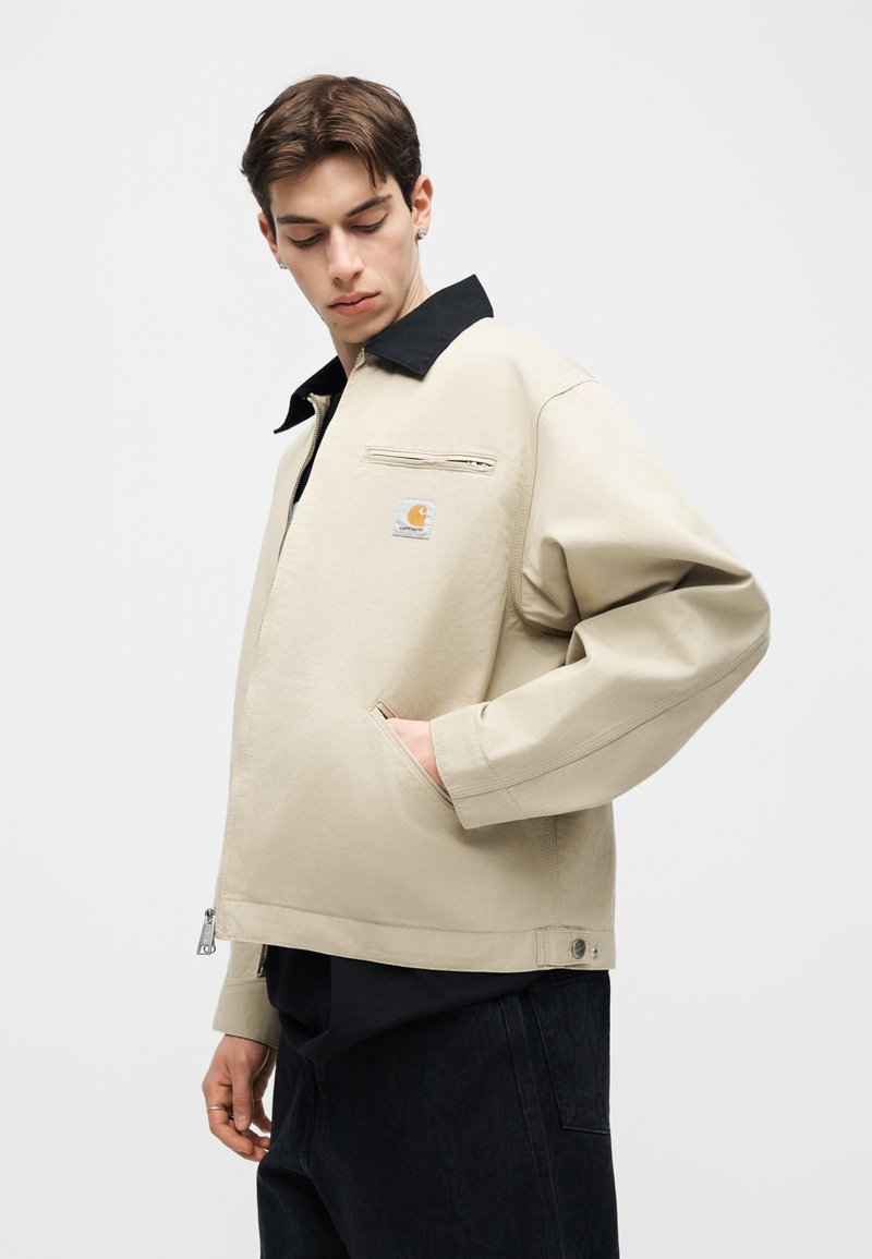 Joven con cabello corto castaño que lleva una chaqueta beige con cuello negro y parche con logo, con las manos en los bolsillos, sobre un fondo claro y liso.