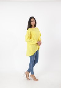 Gelber Strickpullover mit lockerer Passform, langen Ärmeln und rundem Ausschnitt, kombiniert mit blauen Skinny-Jeans und klaren High Heels.