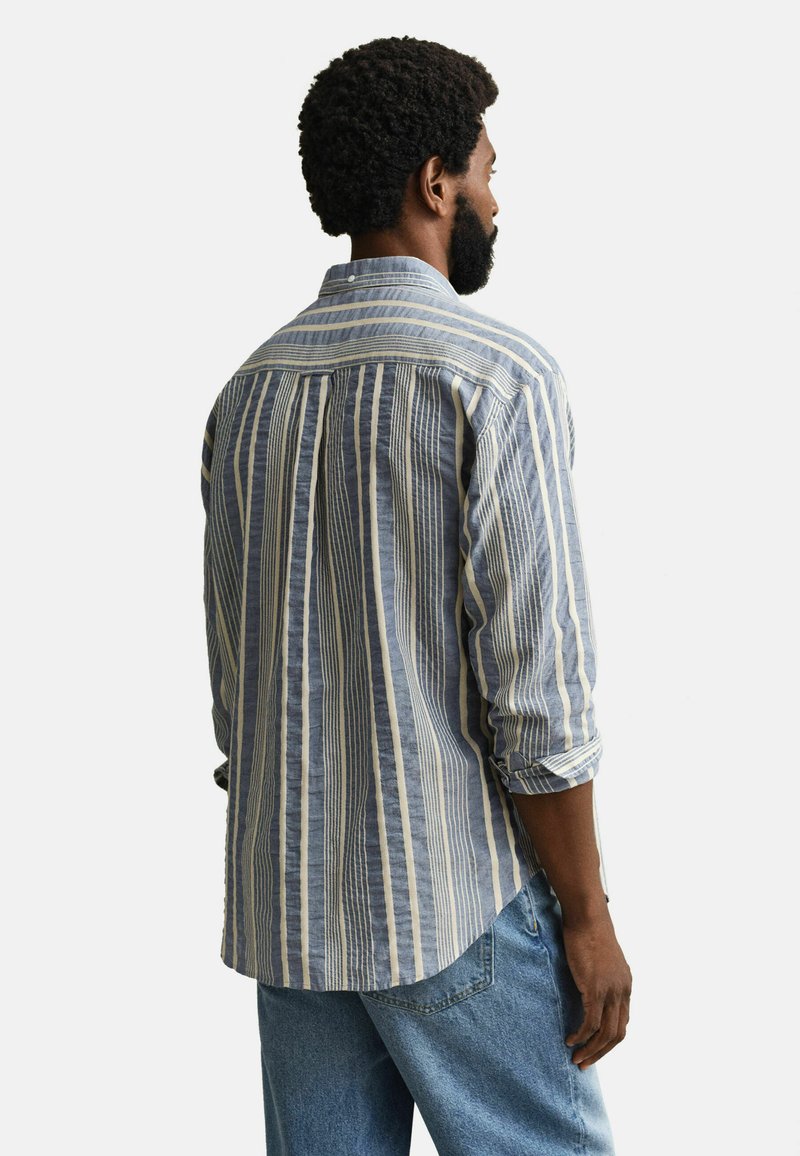 Mann mit lockigem Haar und Bart, der ein blau-weiß gestreiftes Hemd und hellblaue Jeans trägt, mit dem Rücken zur Kamera.