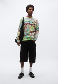 Πολύχρωμο πλεκτό πουλόβερ με γραφικά σχέδια και το κείμενο «UNBREAKABLE», σε συνδυασμό με μαύρο σορτς, λευκές κάλτσες και μαύρα μοκασίνια. Μαύρη τσάντα κρεμασμένη στον ώμο.