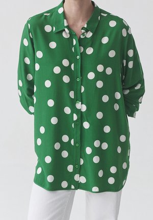 Chemise verte à manches longues avec de gros pois blancs portée avec un pantalon blanc, montrée du cou au milieu de la cuisse.