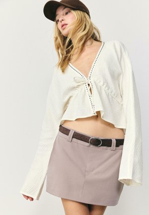 Młoda kobieta ubrana w białą, długorękawową, krótko skrojoną bluzkę z wiązaniem z przodu, taupe mini spódnicę z ciemnym paskiem oraz ciemnobrązową czapkę, pozuje na tle jednolitego tła.