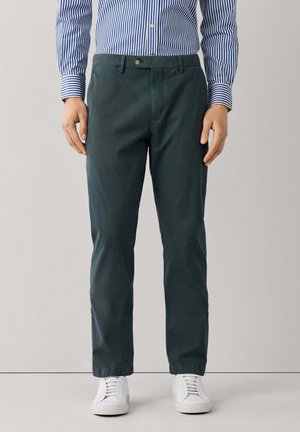 Hombre con pantalones rectos de color verde oscuro, zapatillas blancas y una camisa de manga larga a rayas azul y blanca, de pie frente a un fondo liso.