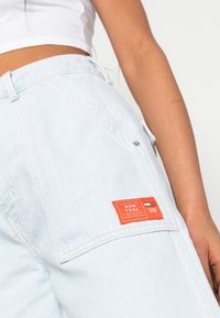Pantalones cargo de denim azul claro con grandes bolsillos laterales, una etiqueta naranja que detalla "NUEVA YORK" y "TOMMY JEANS", con un corte relajado y cordón.