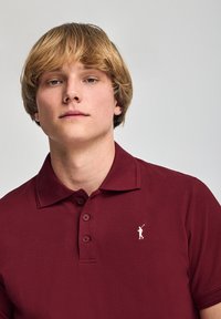 Polo de color rojo oscuro en una tela texturizada, con cuello y tres botones, y un pequeño logo blanco de un golfista en el lado izquierdo del pecho.