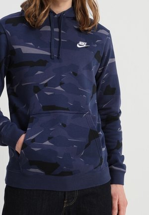 Sudadera azul marino con un patrón de camuflaje en tonos variados, que presenta un bolsillo tipo canguro y un logo de Nike blanco en el pecho.