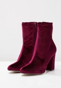 Burgundské sametové kotníkové boty s špičatou špičkou a masivním vysokým podpatkem. Obsahují boční zip pro snadné obutí. Hladká textura po celém povrchu.