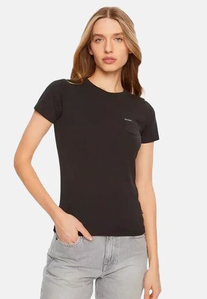 Giovane donna con lunghi capelli biondi che indossa una maglietta nera semplice a maniche corte e jeans grigio chiaro, in piedi con una mano in tasca.