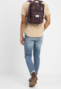 Mężczyzna ubrany w beżową koszulę, niebieskie dżinsy, brązowe buty i niosący ciemnofioletowy plecak, odchodzi na jednolity jasny tle.