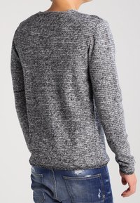 Šedý a černý melírovaný pletený svetr s dlouhými rukávy, kulatým výstřihem a žebrovaným lemem. Není nošený na modrých potrhaných džínech.